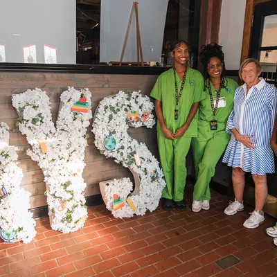 Four women standing next to large floral letters spelling AYS with overlaid educational-themed decorative icons.