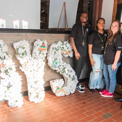 Four smiling people in black shirts pose beside large white floral letters spelling AYS indoors.