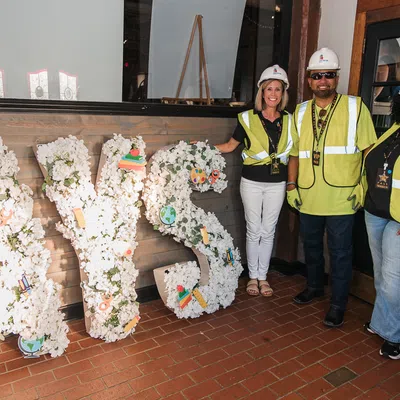Three construction workers in hard hats and reflective vests stand beside large floral letters spelling AVS indoors