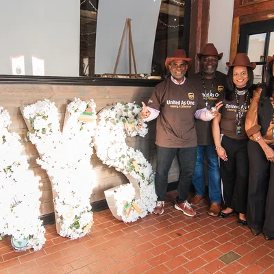 Four people in brown shirts and hats stand beside large floral letters spelling AYS indoors.