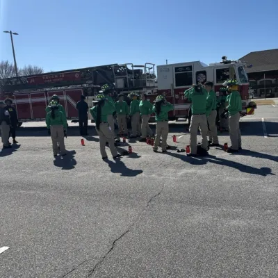 Group of firefighters in green uniforms standing near a red fire truck outdoors on a sunny day.