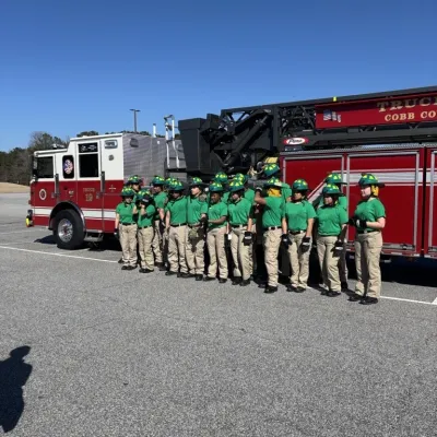 Group of young firefighters in green shirts and helmets standing in front of a red fire truck on clear day.
