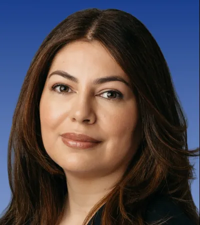 Professional headshot of a woman with brown hair, wearing a black blazer against a blue gradient background.
