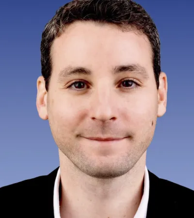 Close-up of a smiling man with short brown hair wearing a black jacket and white shirt on blue gradient background