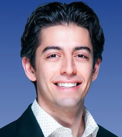 Smiling young man in a dark blazer and white patterned shirt against a blue gradient background