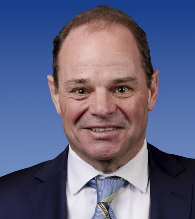 Portrait of a smiling middle-aged man in a dark suit, white shirt, and striped blue and yellow tie against a blue background
