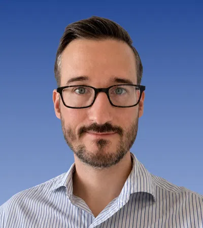 Man with glasses and short beard wearing a striped shirt against a blue gradient background.