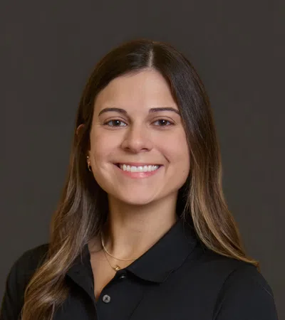 Smiling woman with long brown hair wearing black Pinnacle Orthopaedics polo shirt against dark background.