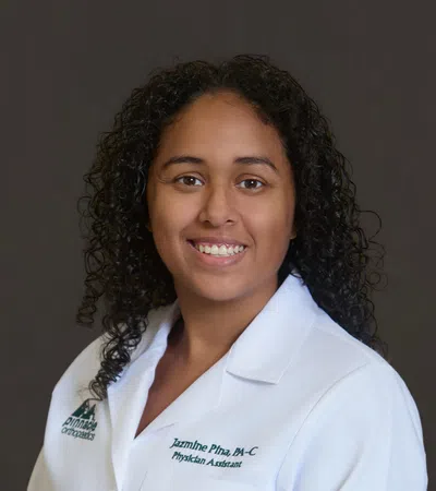 Smiling female physician assistant with curly hair wearing a white medical coat against a dark background