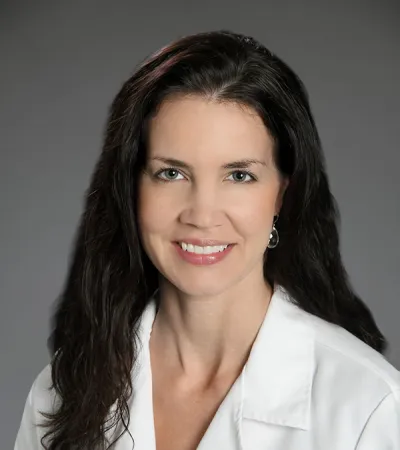 Professional female physician assistant with dark hair wearing white lab coat smiling against gray background.