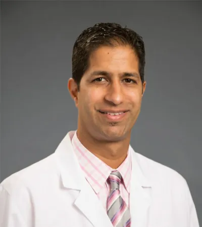 Smiling male doctor in white coat with Pinnacle Orthopaedics logo and striped tie against gray background