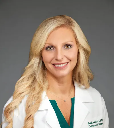 Smiling female doctor with long blonde hair wearing a white Pinnacle Orthopaedics coat and green scrub top.