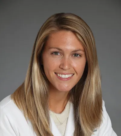 Smiling female doctor with long blonde hair in white coat against a gray background.