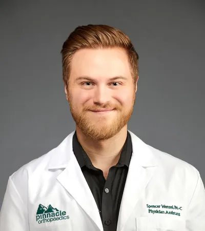 Smiling male physician assistant in white coat with Pinnacle Orthopaedics logo on gray background.
