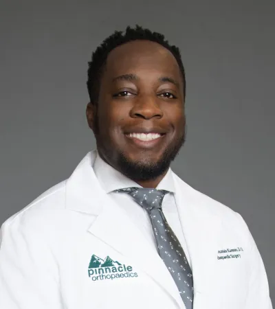 Smiling male orthopaedic surgeon wearing a white coat and a patterned tie against a gray background.