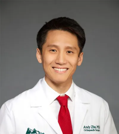 Smiling orthopedic surgeon Andy Zhu MD in white coat and red tie with Pinnacle Orthopaedics logo on grey background