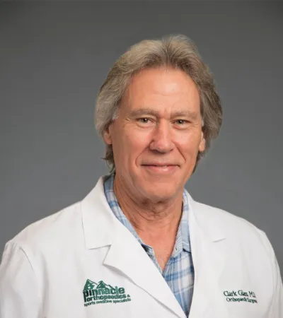 Middle-aged male doctor wearing white coat with Pinnacle Orthopaedics logo and plaid shirt, smiling