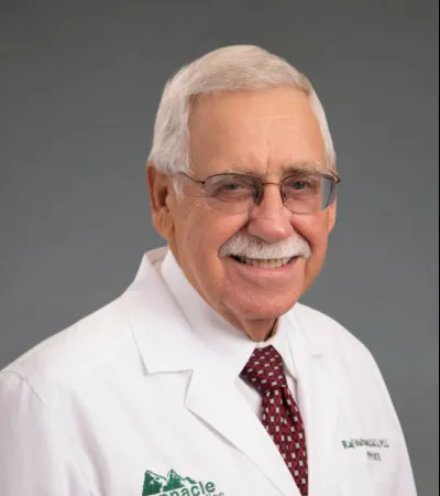 Smiling elderly male doctor wearing glasses and a white Pinnacle Orthopaedics lab coat with a patterned tie.