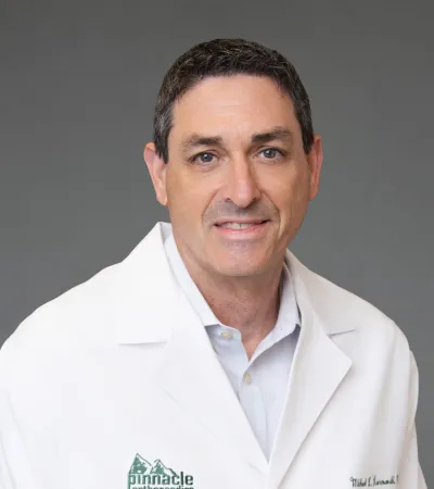 Smiling male doctor wearing a white coat with Pinnacle Orthopaedics logo on a gray background.