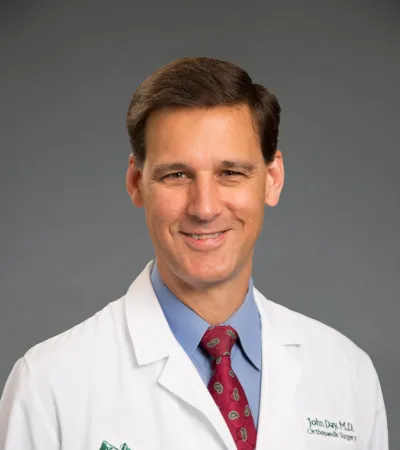 Smiling male doctor in white coat with Pinnacle Orthopaedics logo and red patterned tie on gray background