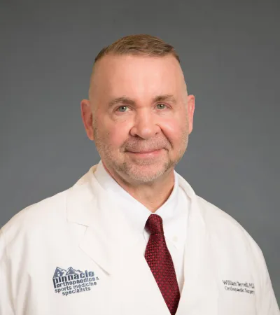 Middle-aged male doctor wearing a white coat with Pinnacle Orthopaedics logo and red tie, smiling against gray background
