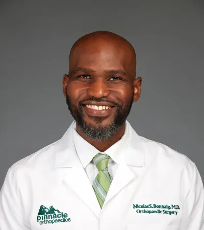 Smiling male doctor wearing white coat and green striped tie with Pinnacle Orthopaedics logo on gray background.