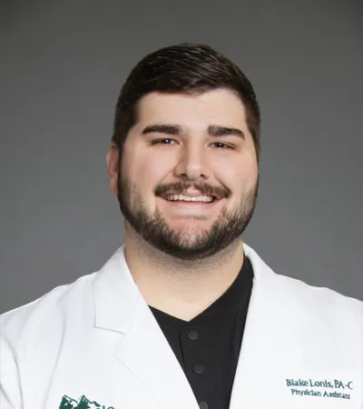Smiling male physician assistant wearing a white medical coat with Pinnacle Orthopaedics logo on gray background.