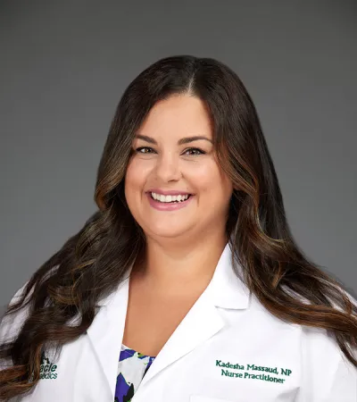 Smiling nurse practitioner with long dark hair wearing white medical coat against gray background.