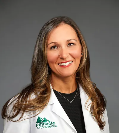 Smiling female doctor wearing a white Pinnacle Orthopaedics lab coat against a gray background.