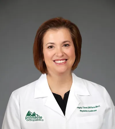 Smiling female physician assistant in white lab coat with Pinnacle Orthopaedics logo on gray background.