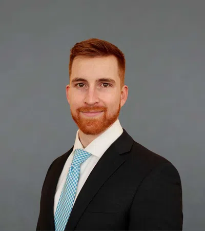 Professional red-haired man in a suit and blue tie smiling against a plain gray background.