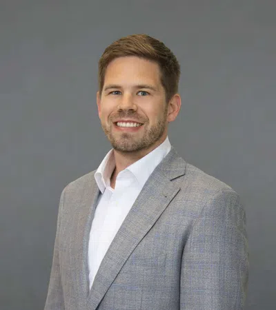 Smiling young man wearing a gray plaid blazer and white shirt against a plain gray background.