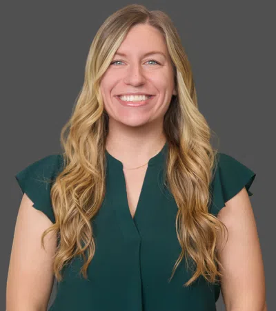 Smiling woman with long blonde hair wearing a dark green blouse against a gray background