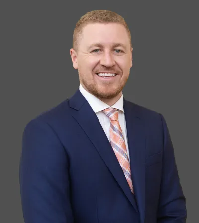 Smiling man in a navy blue suit and orange plaid tie against a solid gray background.