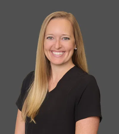 Smiling woman with long blonde hair wearing black scrubs against a plain gray background.
