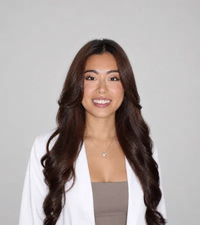 Smiling woman with long dark wavy hair wearing a white blazer and beige top against a plain background