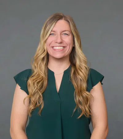 Smiling woman with long blonde hair wearing a green sleeveless blouse standing against a plain gray background