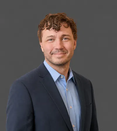 Smiling man with curly hair wearing a dark blazer and light blue shirt against a gray background