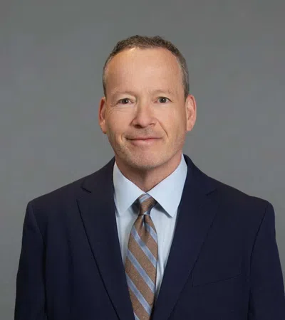 Professional middle-aged man in navy suit and striped tie smiling against a plain gray background