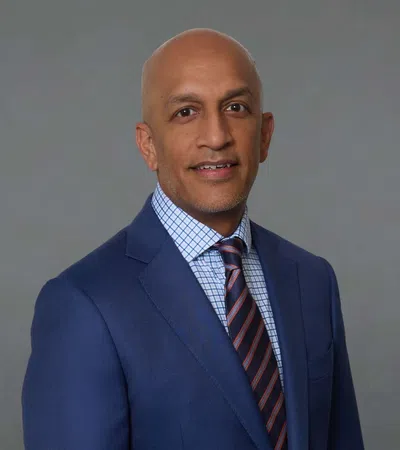 Professional portrait of a bald man in a blue suit, checkered shirt, and striped tie against a gray background.