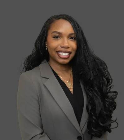 Smiling woman with long curly hair wearing a gray blazer and layered gold necklaces on gray background