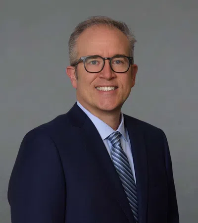 Smiling middle-aged man with glasses wearing a navy suit and striped tie against a plain gray background