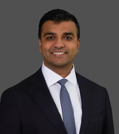 Professional man in dark suit and patterned tie smiling against a solid gray background.