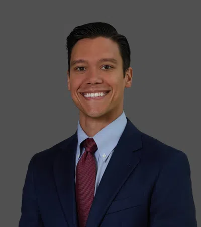 Smiling young man in navy suit, light blue shirt, and red tie against a gray background.