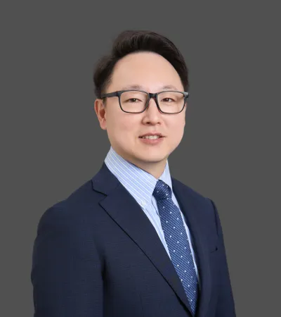 Professional man wearing glasses, dark suit, striped shirt, and patterned tie on gray background.