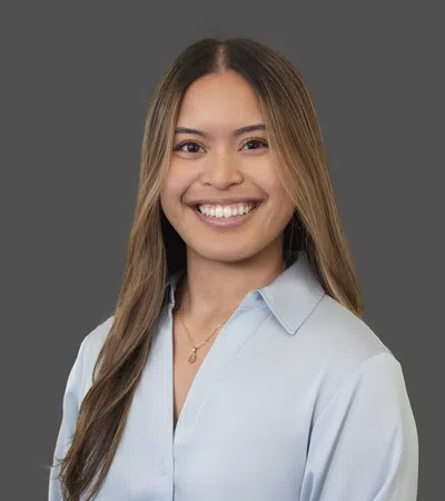 Smiling young woman with long brown hair wearing a light blue blouse and gold necklace on gray background
