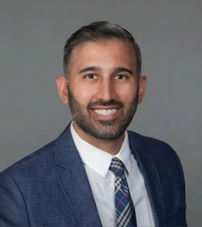 Smiling man with beard wearing a blue suit and plaid tie against a gray background.