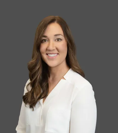 Smiling woman with long brown hair wearing a white blouse against a plain gray background.