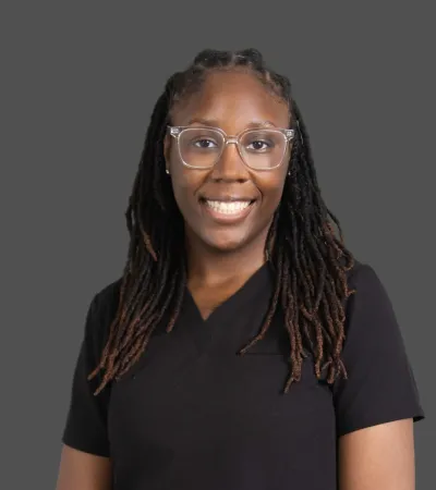 Smiling woman with dreadlocks and clear glasses wearing a black top against a gray background