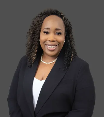 Smiling professional Black woman with curly hair wearing a black blazer, white blouse, and pearl necklace on gray background